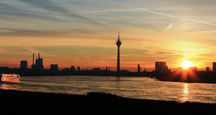 Düsseldorf skyline at sunset with Rhine Tower, bridges and glowing orange sky reflected on the river.