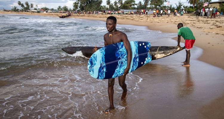 Young man carrying a blue surfboard along a lively beach shoreline.
