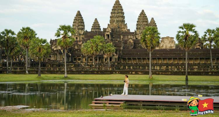Lone visitor stands on a wooden platform before the vast facade of Angkor Wat across a moat.