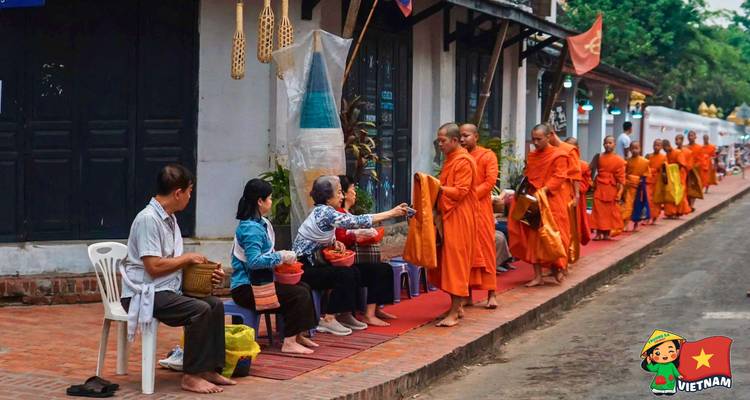 Line of saffron-robed monks receives alms from locals seated on a brick walkway.