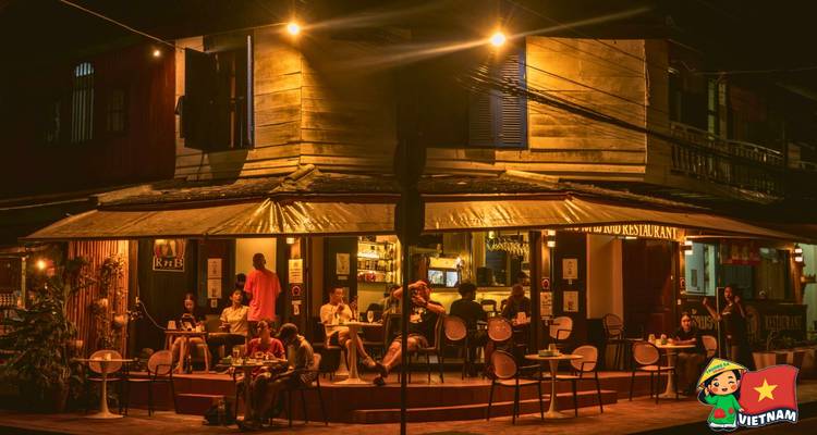 Warmly lit street-corner café buzzes with patrons at wooden tables on a tropical night.