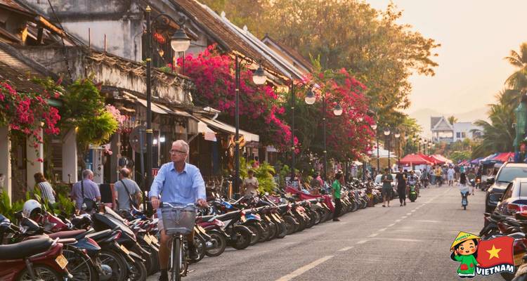 Bustling flower-lined street filled with scooters, shops and cyclists in late afternoon light.