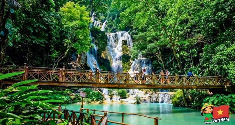 Visitors cross a wooden bridge in front of cascading turquoise waterfalls set in dense jungle.