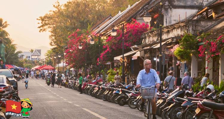 Bustling flower-lined street filled with scooters, shops and cyclists in late afternoon light.