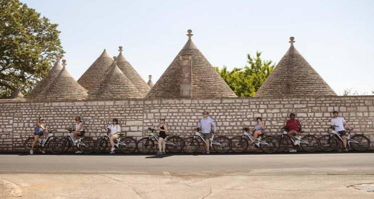 Rangée de cyclistes se reposant contre un mur en pierre surmonté de trulli sous un ciel dégagé