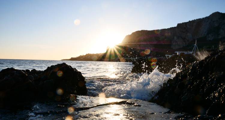 Low-angle shot of waves splashing against dark rocks at sunset with sunrays flaring over a coastal cliff.