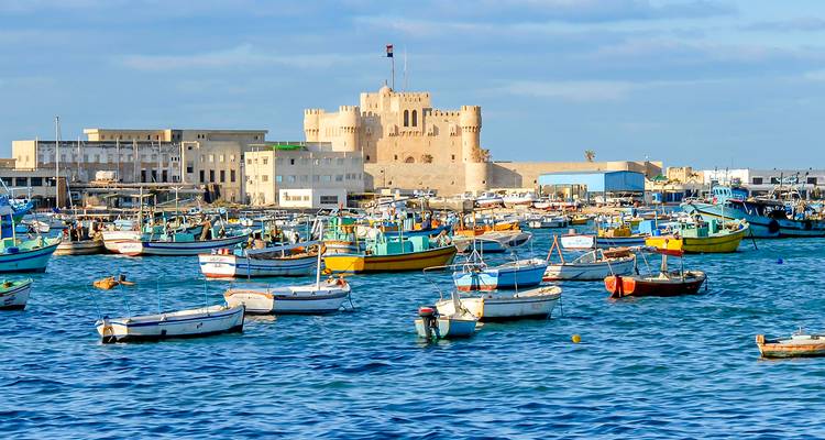 Colorful fishing boats float in a busy harbor with a historic stone fortress on the waterfront