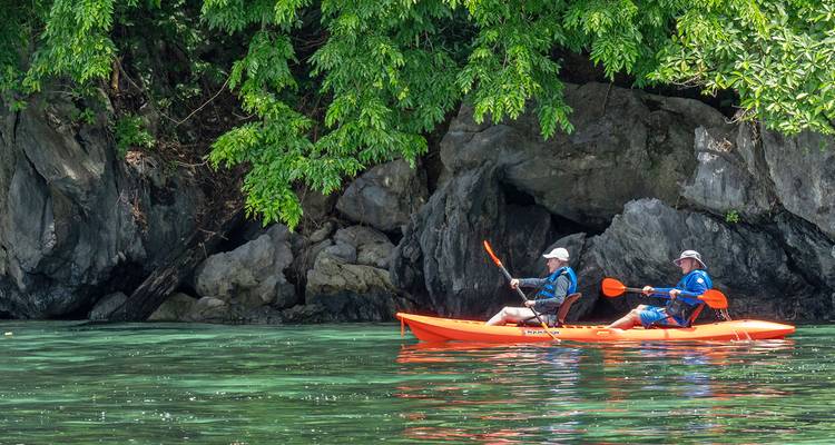Two kayakers paddle an orange kayak on crystal-clear turquoise water beneath lush limestone cliffs and jungle foliage.