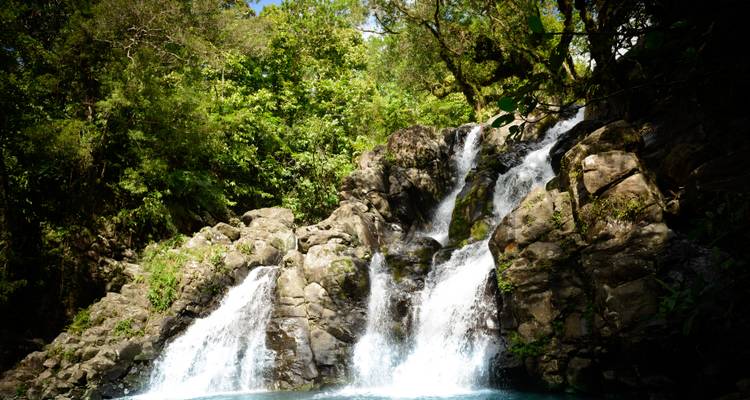Multi-tiered tropical waterfall cascading over dark rocks into a blue pool surrounded by dense jungle.