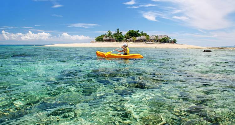 Kayaker paddles across crystal-clear turquoise water toward a tiny sandy island with palm trees under bright blue sky.