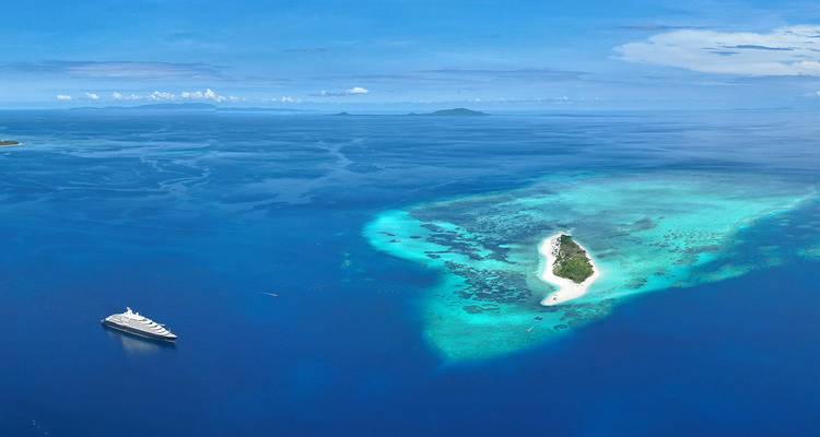 Aerial view of small coral island and luxury yacht amid vivid blue Indian Ocean.