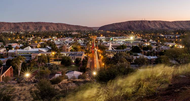 Dusk aerial view over Alice Springs with glowing streetlights and surrounding red desert ranges.