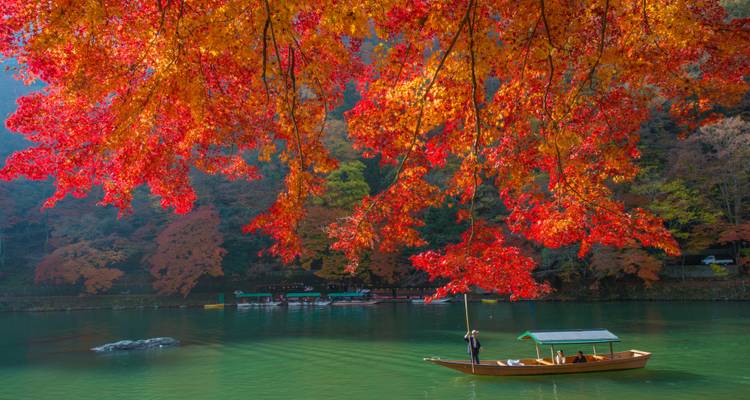 Traditional wooden boat floats on calm river beneath vivid red autumn leaves in Arashiyama