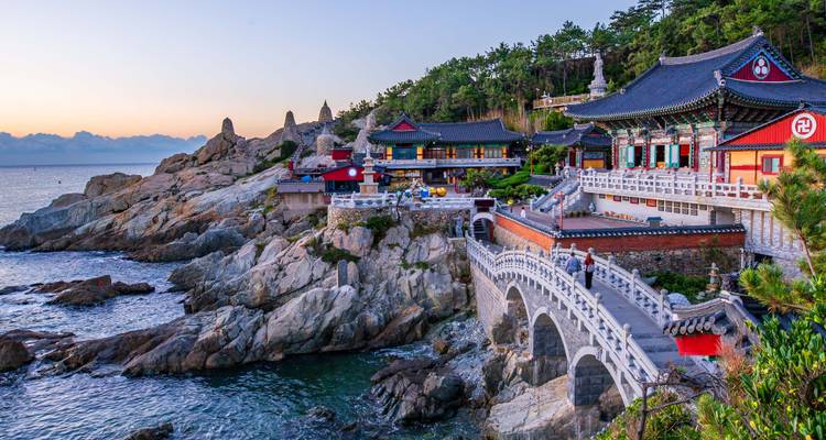 Dramatic seaside temple complex perched on rocky cliffs at sunrise with stone bridge and pagodas.