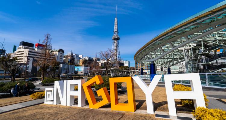Large white and orange @NAGOYA letters in a city plaza with the Nagoya TV Tower and modern skyline under a clear blue sky.