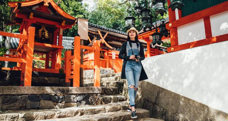 A young woman with a hat and camera walks up stone steps amid vivid red shrine gates and lanterns in a lush temple courtyard.