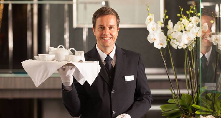 Un serveur souriant en costume sombre tenant un plateau de thé devant un décor élégant