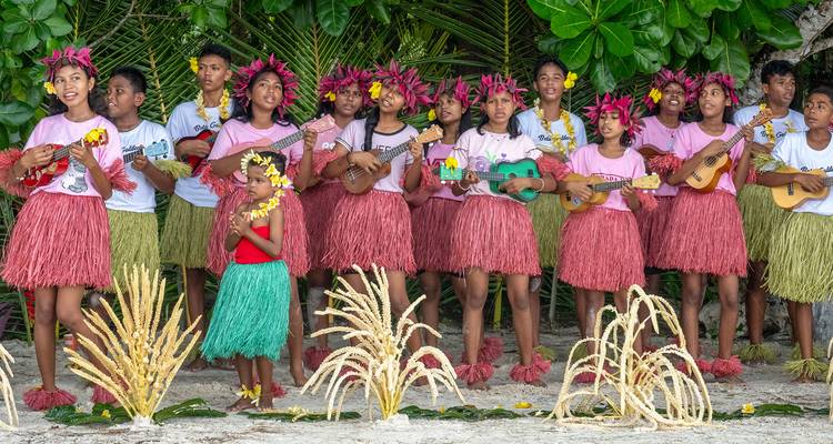 Island youth group wearing flower crowns and grass skirts perform music with ukuleles on a sandy beach