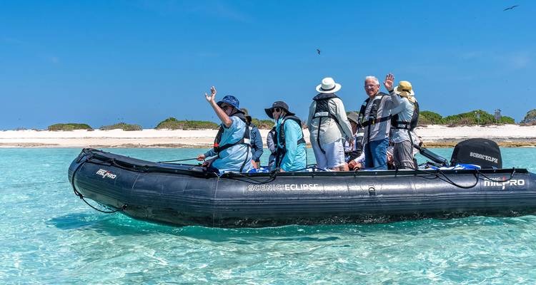 Tourists in life-jackets waving from a black inflatable zodiac on crystal-clear turquoise water near sandy shore.