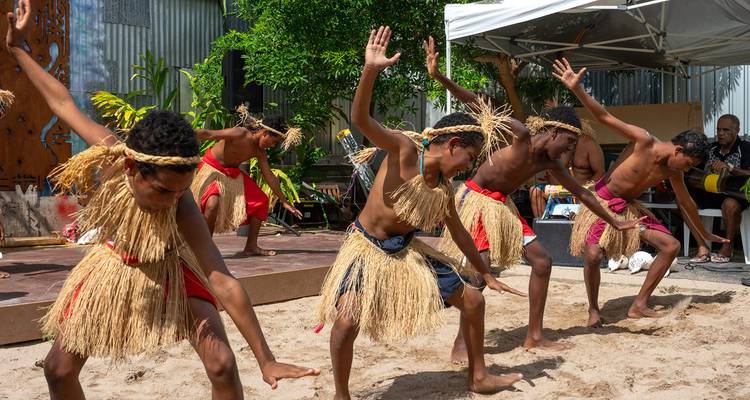 Young dancers in traditional grass skirts perform an energetic cultural dance outdoors on a sandy stage.
