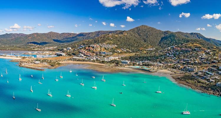 Panoramic aerial of turquoise bay dotted with sailboats and hillside town at Airlie Beach.