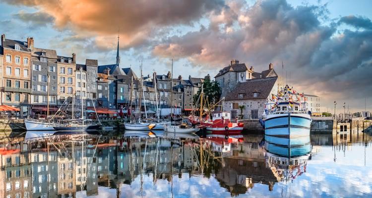 Port pittoresque d'Honfleur avec des bateaux colorés, des bâtiments historiques et des nuages de coucher de soleil spectaculaires reflétés sur une eau calme.