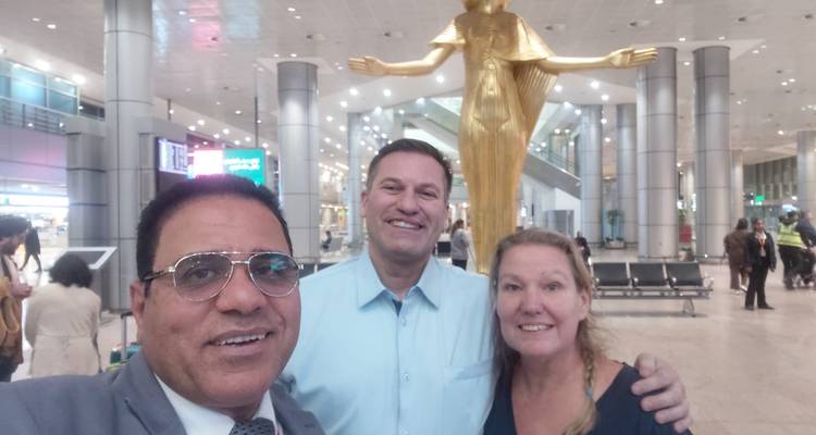 Le guide pose avec un couple souriant à l'aéroport du Caire sous une statue dorée.