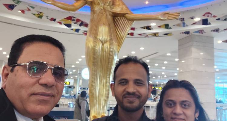 Trois voyageurs posent pour un selfie dans le terminal d'un aéroport devant une statue de style pharaon doré et des luminaires de plafond brillants.