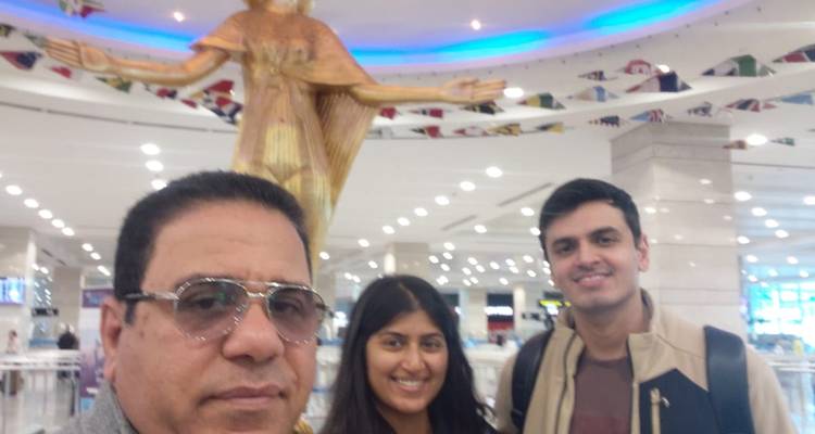 Selfie de trois personnes souriantes à l'intérieur d'un aéroport avec une statue dorée en arrière-plan, légèrement floue.