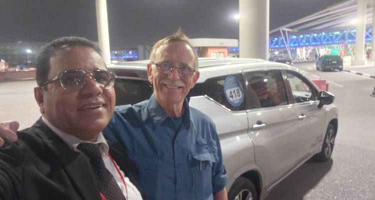 Deux hommes sourient pour un selfie à côté d'une camionnette devant un aéroport la nuit sous un éclairage artificiel.