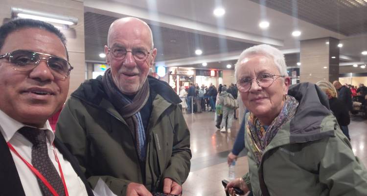 Trois voyageurs expérimentés se tiennent ensemble dans un hall d'arrivée en tenant des documents, entourés par d'autres passagers.