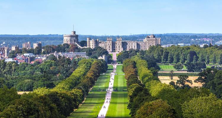 View down a long, tree-lined avenue leading to Windsor Castle under a bright blue sky, with visitors walking along the path.