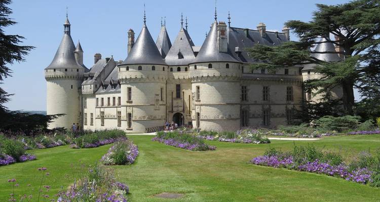 Fairytale-style stone chateau with conical towers, surrounded by colorful flowerbeds and green lawns.