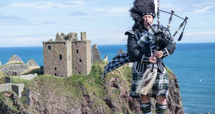 Bagpiper in full Highland dress standing on a cliff with medieval castle ruins and sea behind