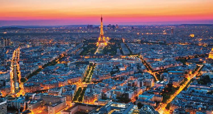 Twilight aerial panorama of Paris with the Eiffel Tower illuminated and city lights glowing against a colorful sunset sky.