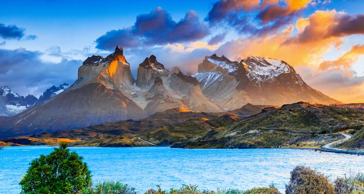 Die dramatischen Gipfel der Torres del Paine leuchten in Sonnenaufgangsfarben über einem blauen See und hügeliger Landschaft.
