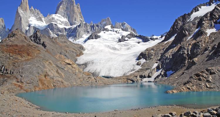 Eine ruhige, türkisfarbene Lagune erstreckt sich unterhalb der schneebedeckten Granitpfeiler des Mount Fitz Roy unter einem klaren Himmel.