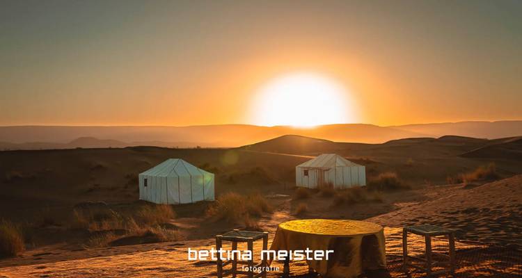 Sonnenaufgang über Wüsten-Glamping-Zelten mit Tisch im Vordergrund, Wasserzeichentext am unteren Rand.