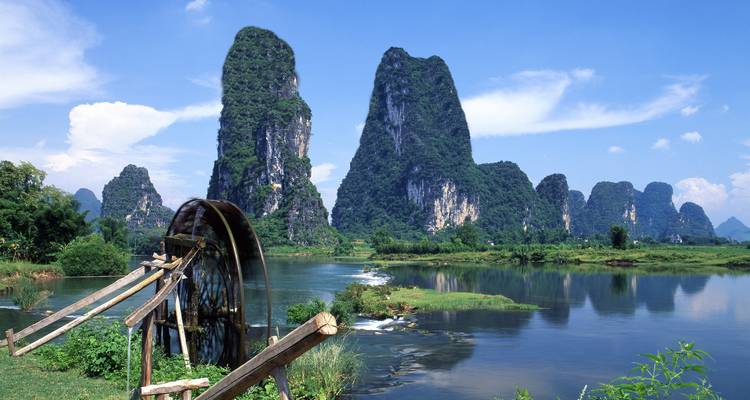 Karstkalkssteinkuppen erheben sich hinter einem hölzernen Wasserrad auf einem ruhigen Fluss im ländlichen China.