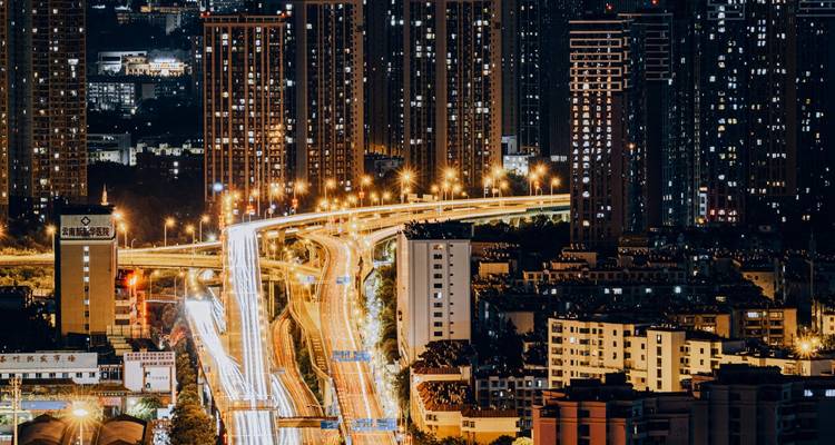 Nighttime cityscape of high-rise buildings with light trails from busy elevated highway.