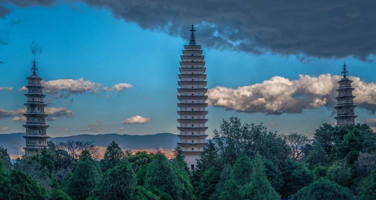 Three ancient pagodas rise above green forests with dramatic clouds overhead.