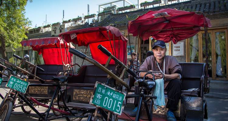 Fahrer einer Rikscha entspannt sich auf seinem dreirädrigen Gefährt unter roten Markisen in einer historischen Hutong-Gasse in Peking