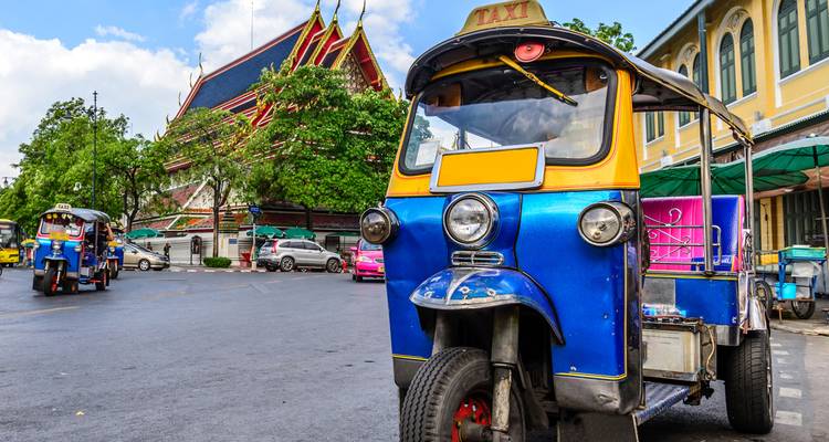 Un tuk-tuk taxi azul brillante estacionado en una calle de la ciudad frente a un colorido templo tailandés bajo un cielo soleado.