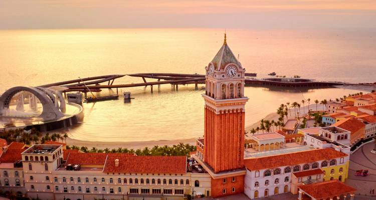 Una campanario de estilo veneciano y edificios costeros en colores pastel brillan con la luz dorada del atardecer junto a una bahía tranquila.