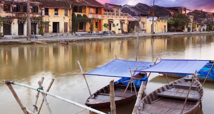 Les maisons-boutiques jaunes traditionnelles se reflètent dans la rivière tranquille de Hoi An au crépuscule, avec des bateaux en bois amarrés au premier plan.