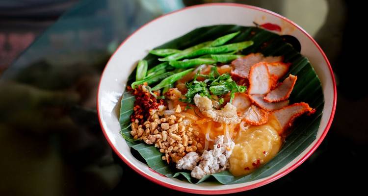 Colourful Thai noodle dish topped with roast pork, peanuts and green beans served on a banana leaf-lined bowl.