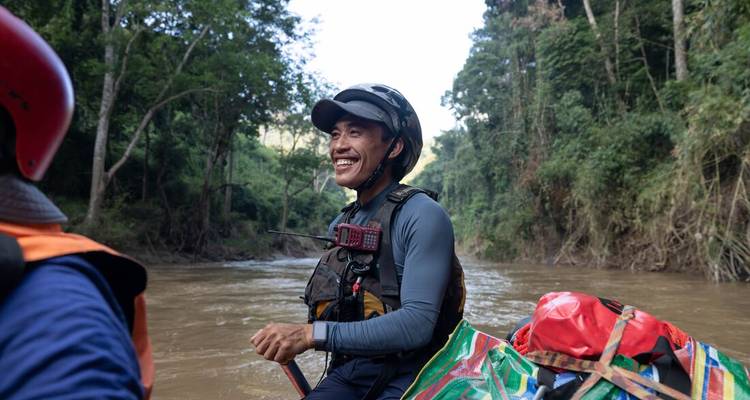 Smiling rafter wearing helmet and life jacket navigates a muddy jungle river with gear bags on the boat.