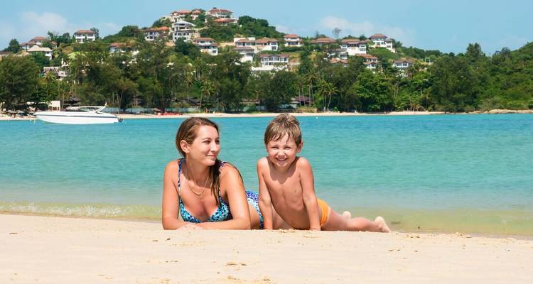 Mother and young son relax on a white-sand beach beside calm turquoise water with hillside villas in the background.
