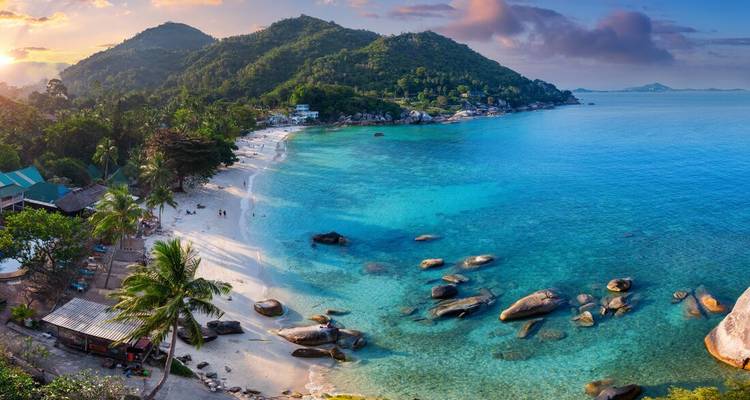 Panoramic aerial view of a tropical island bay with turquoise water, white sand and lush green hills at sunset.