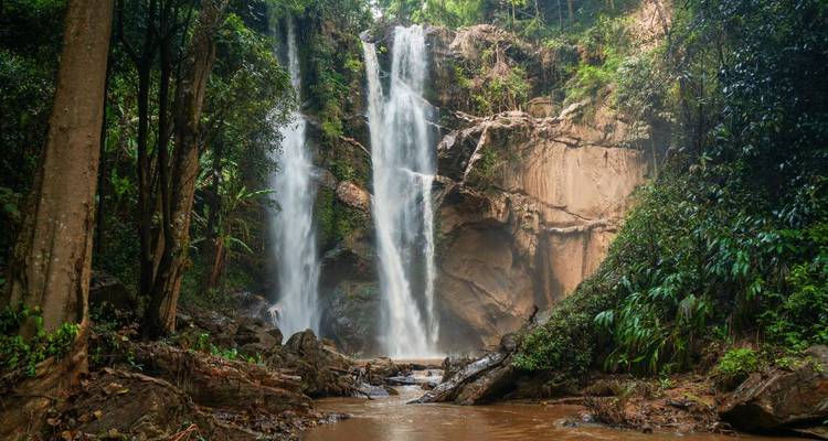 Twin streams of a jungle waterfall cascade into a rocky pool surrounded by dense green vegetation.